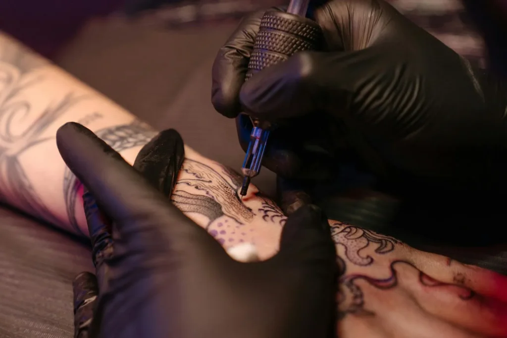 A pair of hands wearing black latex gloves, with the right hand holding a tattoo pen and the left hand holding onto a person's arm while tattooing