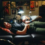 A man wearing a black t-shirt is lying down while getting a tattoo on his left hip Image Filename: Best Tattoo Shops & Studios, Krabi