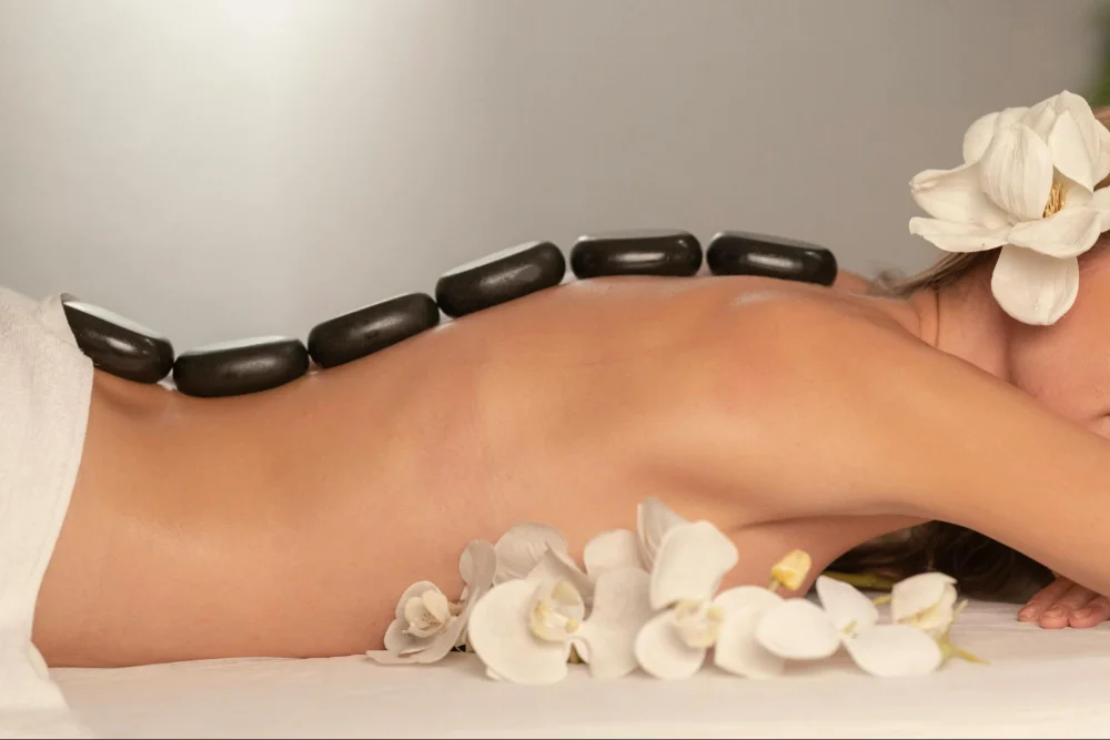 A person receiving a hot stone massage with black basalt stones aligned along their spine, complemented by white orchid flowers placed around their shoulder area.