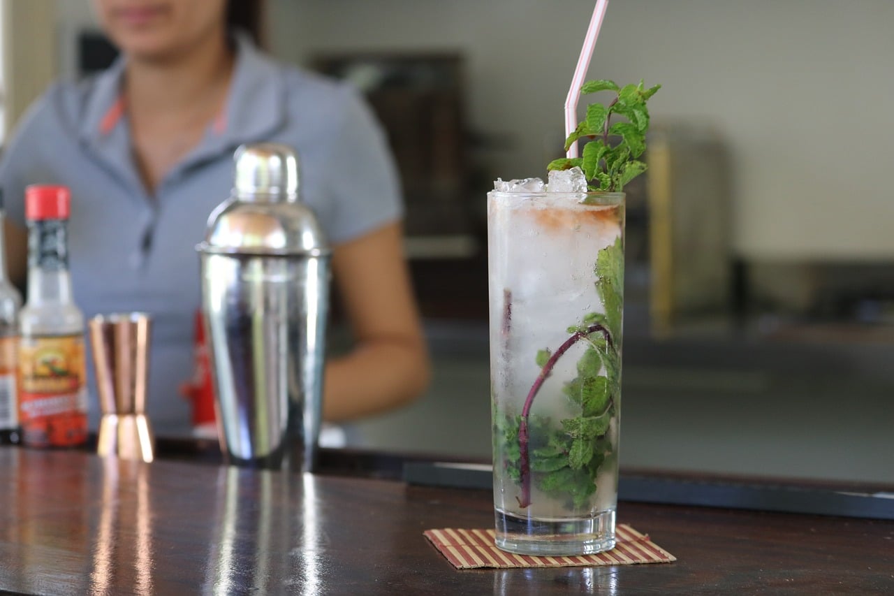 A clear glass with a mojito cocktail drink on top of the bar table served by the female barista.
