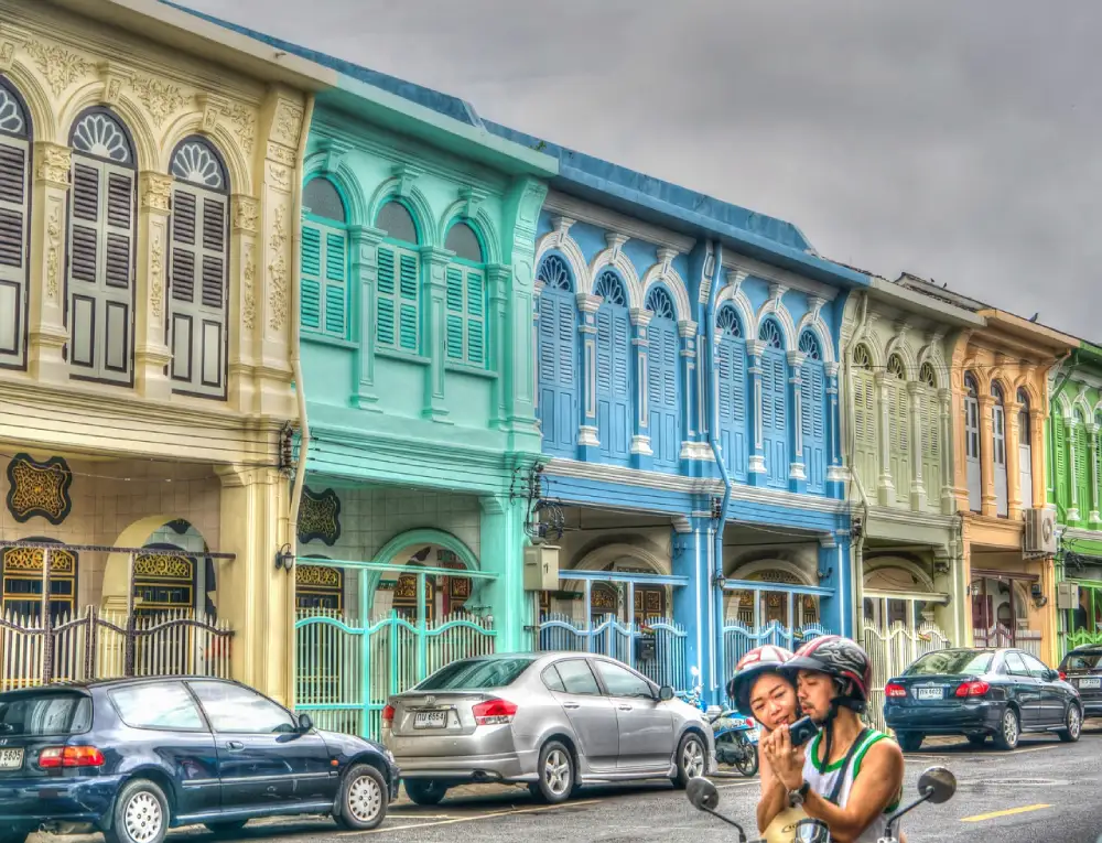 A couple on a scooter in front of a row of colorful, ornate buildings, highlighting a lively street atmosphere.